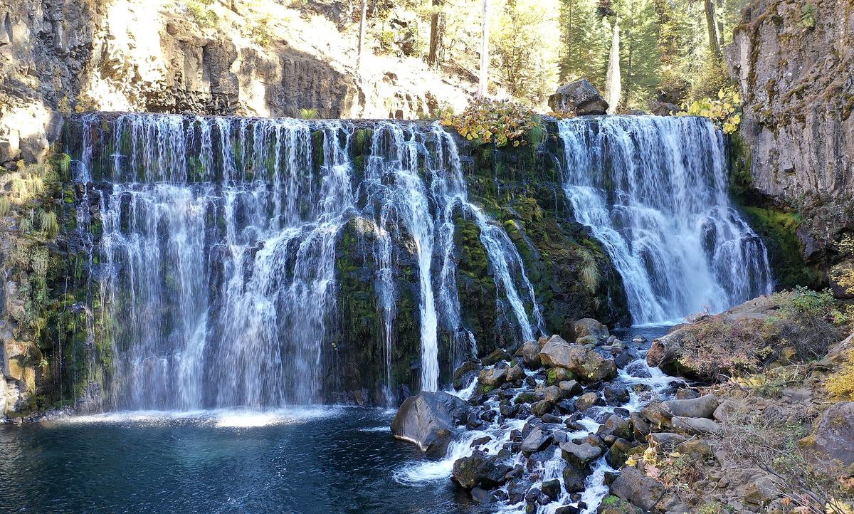 Middle Falls on the McCloud River in autumn, a wide curtain of water pouring over a dark basalt ledge into a blue pool, with yellow leaves along the rim